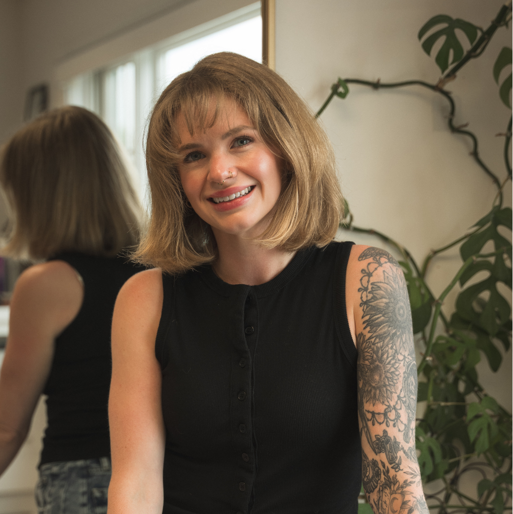 A smiling woman with a shoulder-length blonde bob stands indoors wearing a sleeveless black button-front top and blue jeans. A mirror behind her shows her back, and a leafy plant is visible against a light wall.